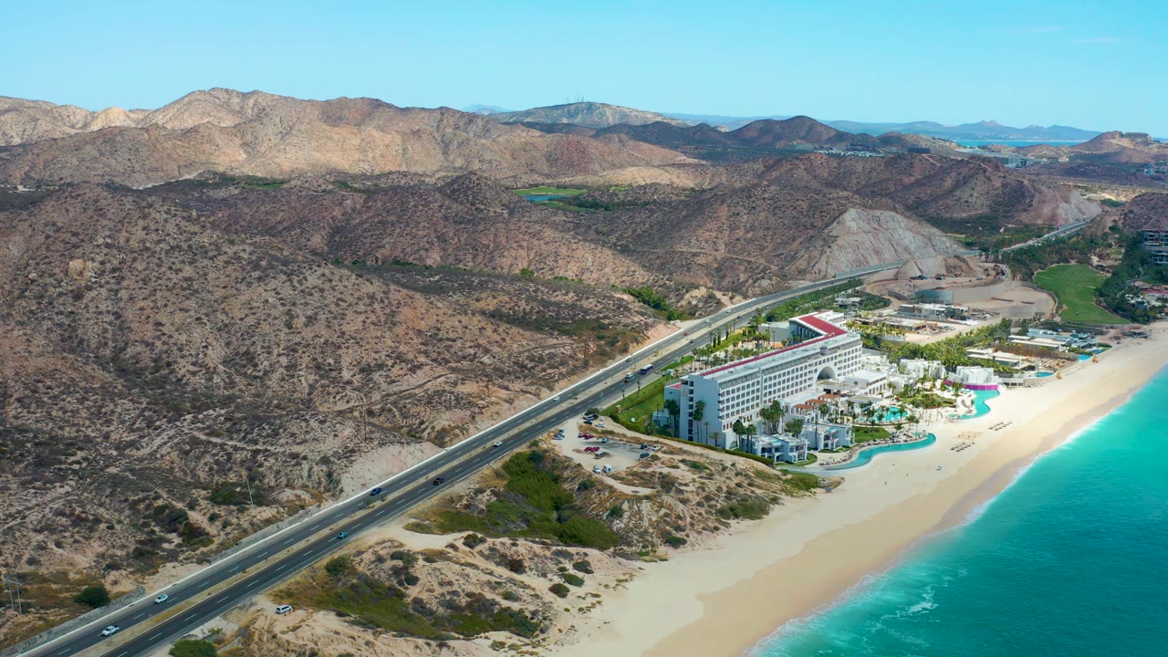 Aerial frame over Marquis Los Cabos with cars spaced along the coastal road, white beachfront structures near the sand, turquoise ocean to the right, rocky hills beyond, and no visible people.