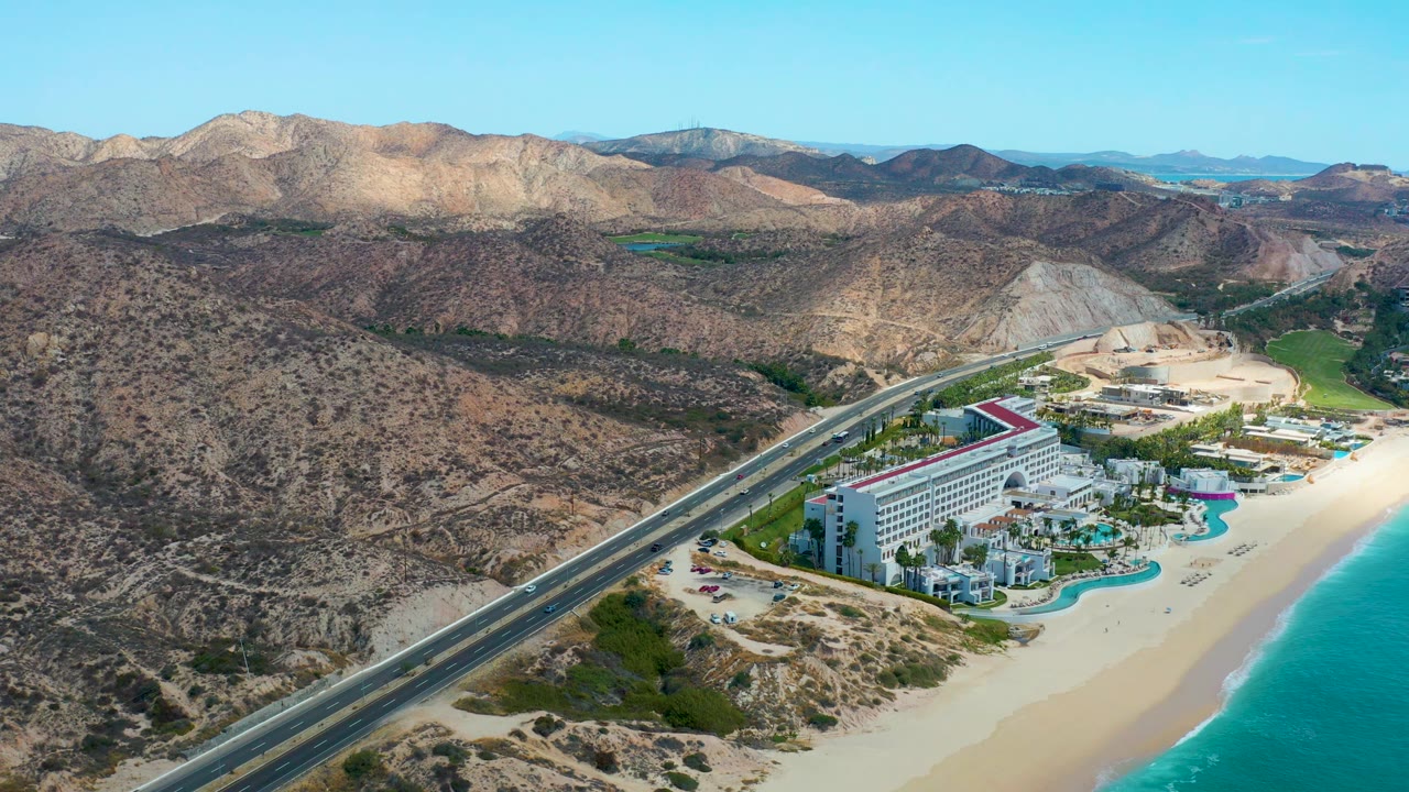 Aerial frame at Marquis Los Cabos, Mexico, showing the hotel lower in frame, the coastal road cutting across the scene, beach and ocean on the right, rugged hills behind, and no visible people.