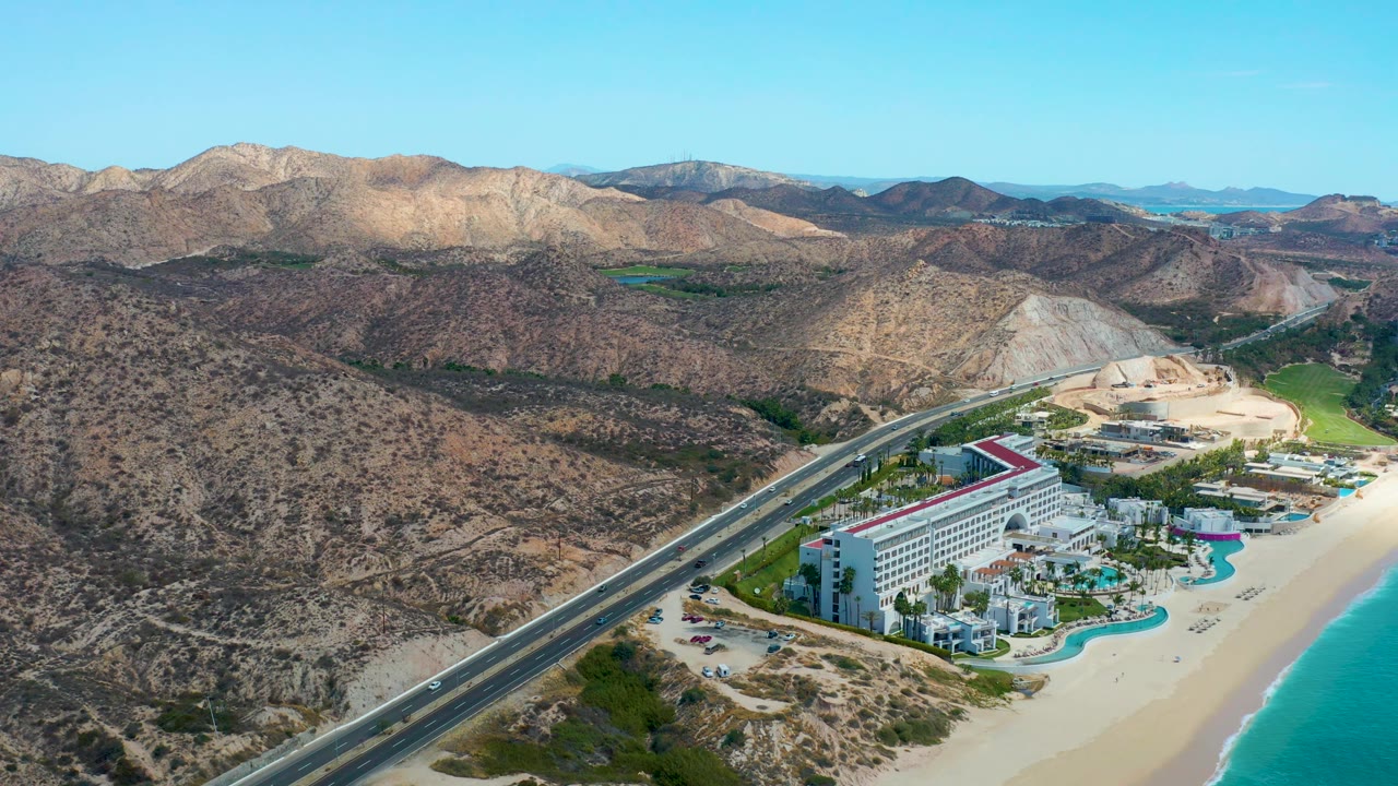 Mid-clip aerial frame at Marquis Los Cabos, Mexico, with the hotel and pool area closer to the lower edge, a diagonal highway, sandy shore, blue ocean strip, dry mountain terrain, and no visible people.