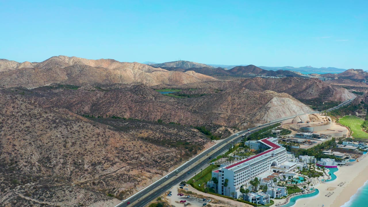 Wide aerial view of coastal resort, road traffic, beach, and mountains.