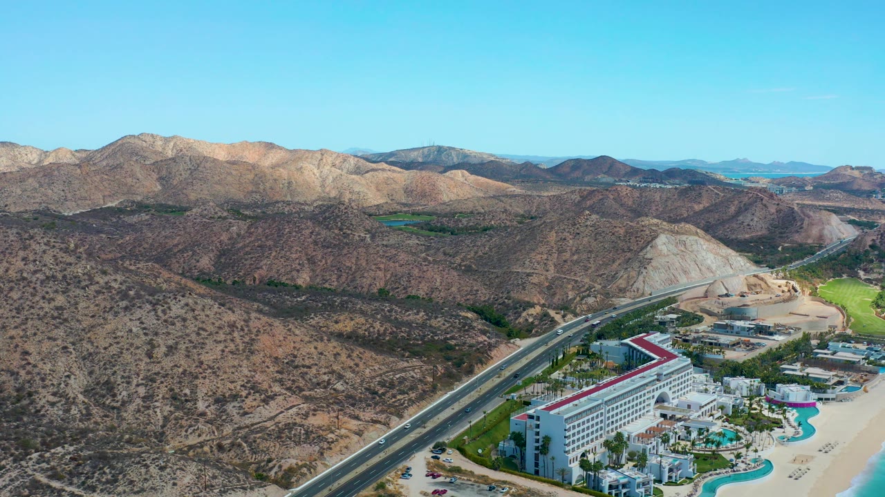 A high aerial frame over Marquis Los Cabos with the resort and pools near the beach, highway traffic at left, rocky hills beyond, distant blue water, and no visible people.