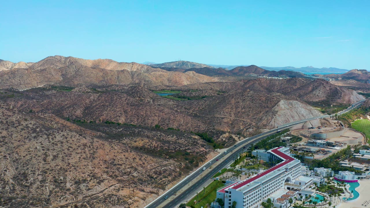 Aerial frame at Marquis Los Cabos, Mexico, showing the highway running up from the lower left past the hotel complex, with brown hills, pale cut slopes, mountains, clear sky, and no visible people.