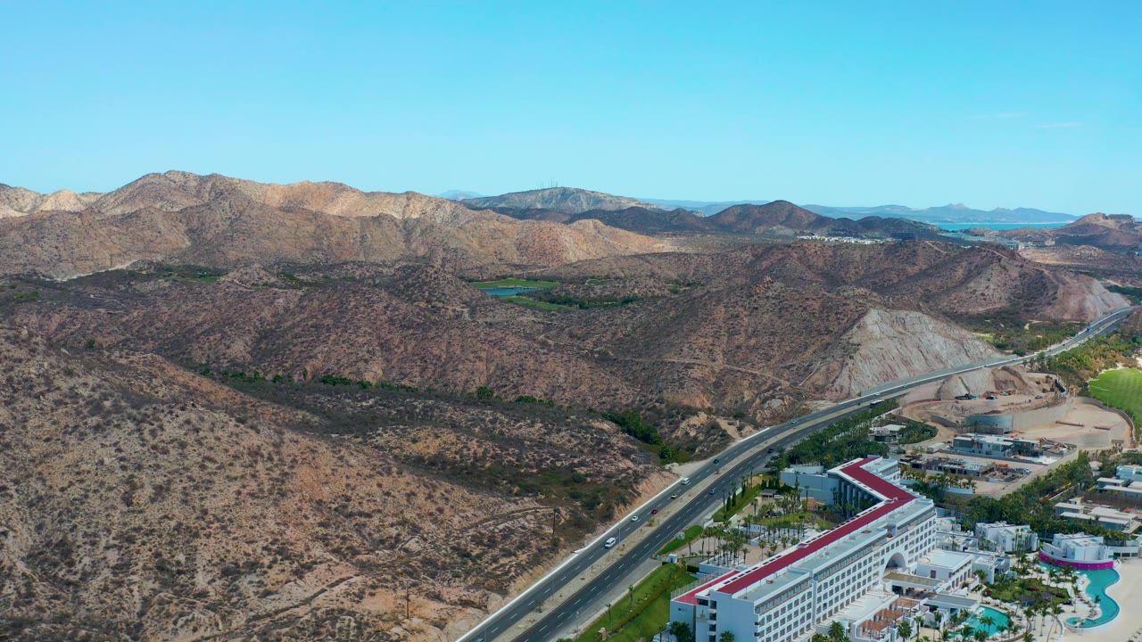 Wide aerial frame from Marquis Los Cabos, Mexico, with the hotel roof clipped along the lower edge, traffic on the highway, layered dry hills, distant mountain ridges, and no visible people.