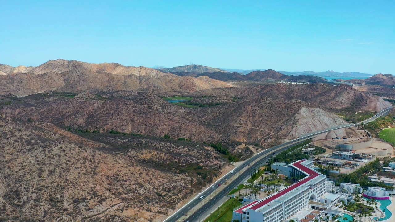 Mid-clip aerial view at Marquis Los Cabos, Mexico, with the highway and small cars cutting past the red-roof hotel and pool area below dry hills and clear blue sky, with no visible people.