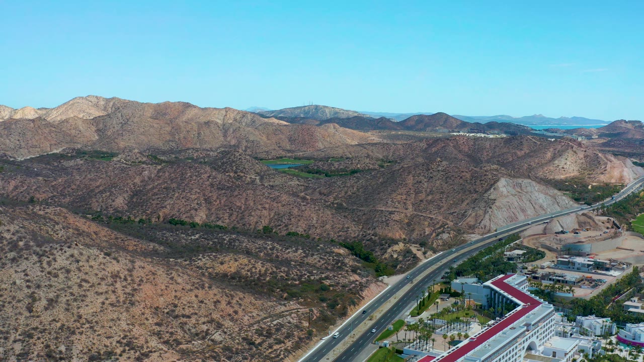 At 0:01, the aerial view keeps the highway lower in frame while rocky hills and pale mountain ridges fill the background at Marquis Los Cabos, with no visible people.