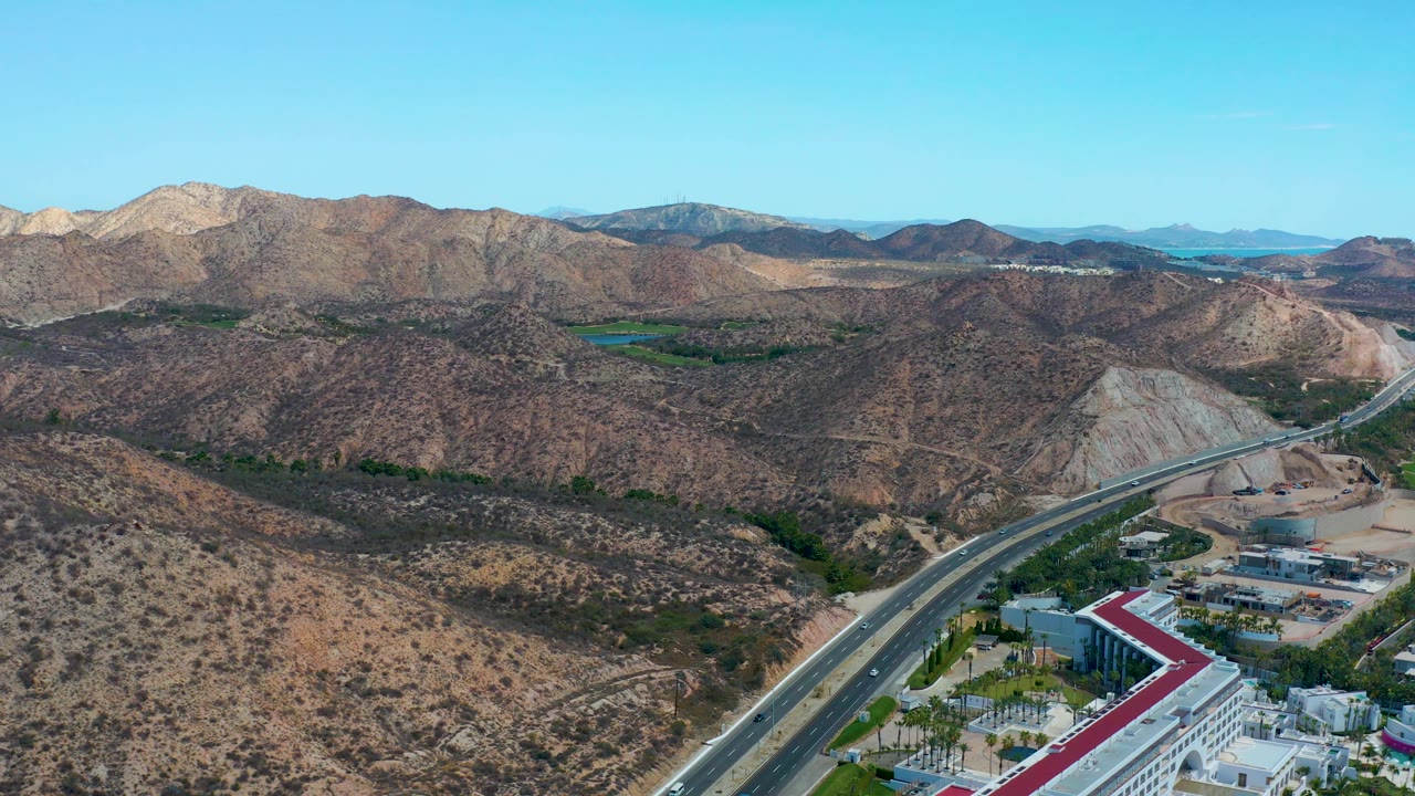 At 0:03, the road bend and building roofs sit low in the frame, with layered rocky hills and tan mountain slopes stretching across Marquis Los Cabos, and no visible people.
