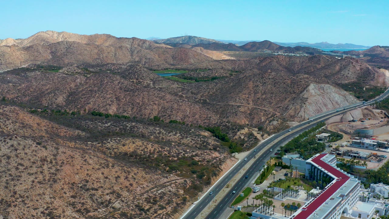 Opening aerial frame over Marquis Los Cabos, Mexico, showing a divided highway, rocky hills, sparse vegetation, a white red-roofed building, distant water, and no visible people.