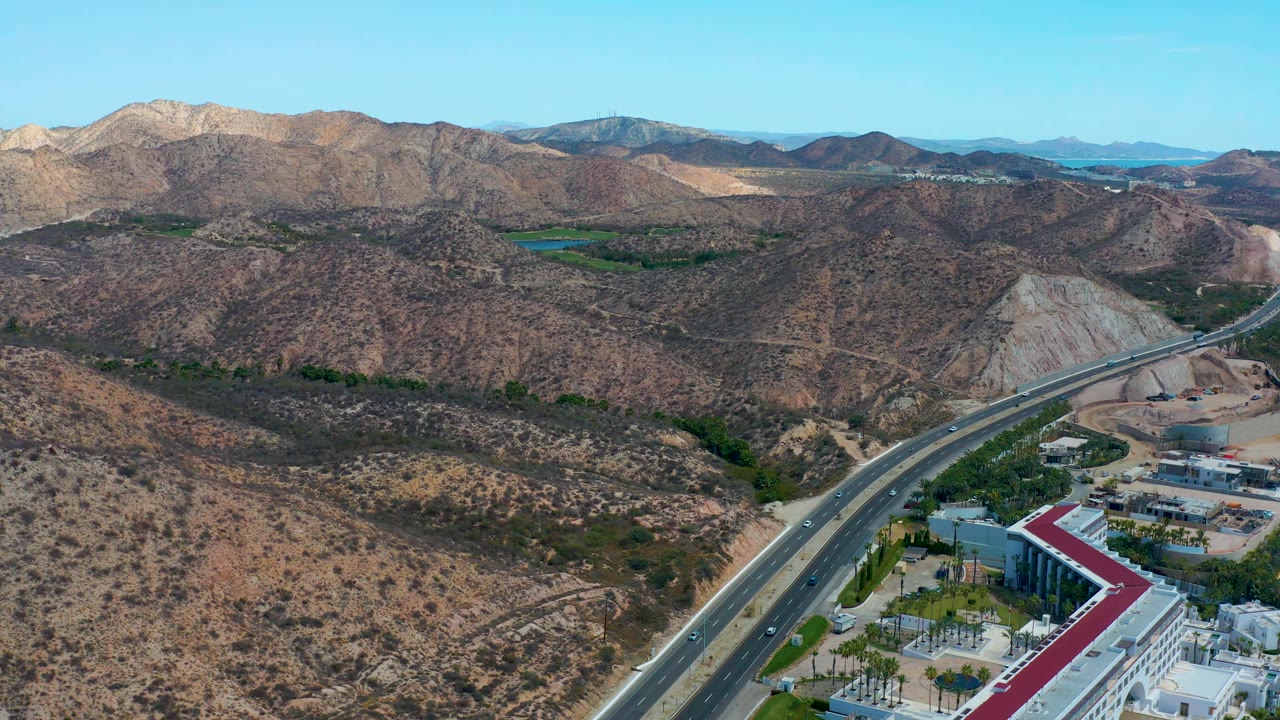 Aerial frame at 0:01 over Marquis Los Cabos, Mexico, with the highway bending through arid hills, small vehicles on the road, green patches in the valley, and no visible people.