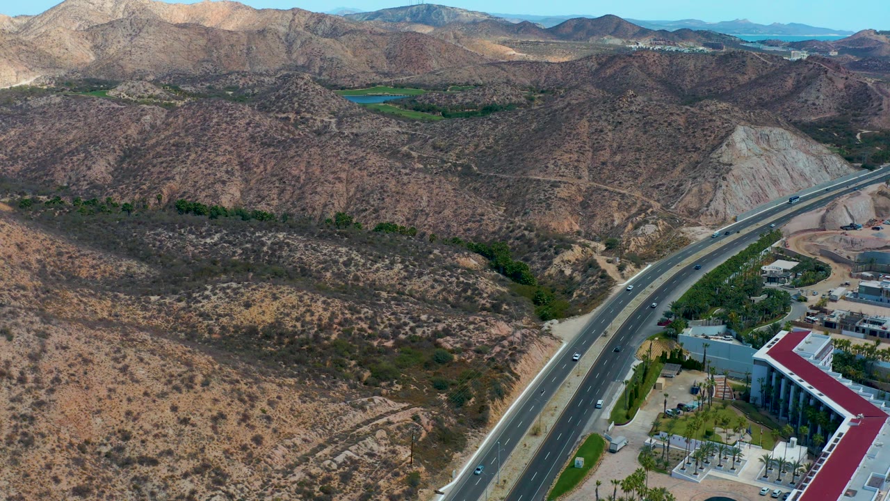 Aerial view in Marquis Los Cabos, Mexico, with cars spaced along the curving highway, scrub-covered hills, pale exposed earth, distant water, and no visible people.