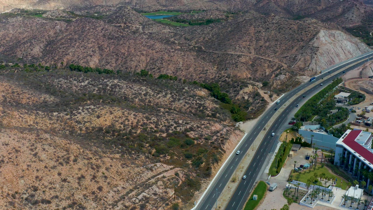 Aerial frame from Marquis Los Cabos, Mexico, showing a divided highway bending through arid hills, small cars on the road, a red-roofed building edge at lower right, and no visible people.