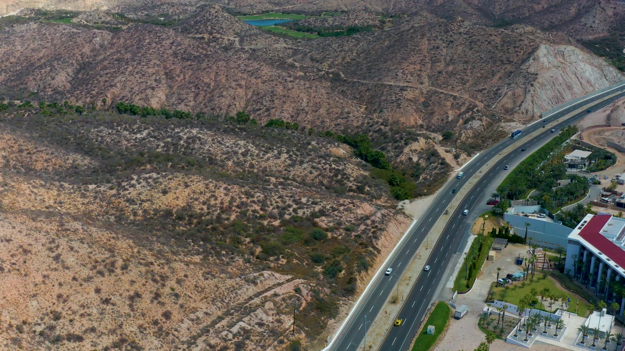 Aerial frame from Marquis Los Cabos, Mexico, showing vehicles spaced along the curved highway, a partial red-roofed building at the lower right, dry hillsides, and no visible people.