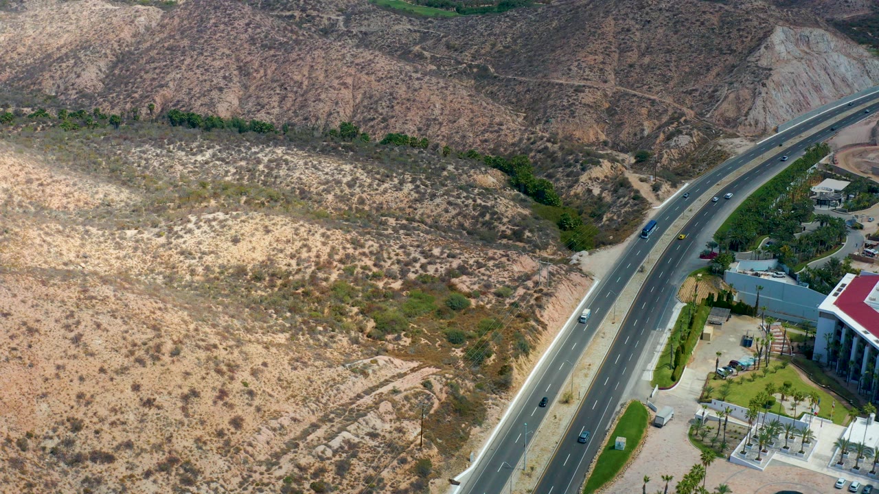 Aerial frame at Marquis Los Cabos, Mexico showing a divided curved highway beside dry scrub-covered hills, vehicles on the road, buildings at lower right, and no visible people.