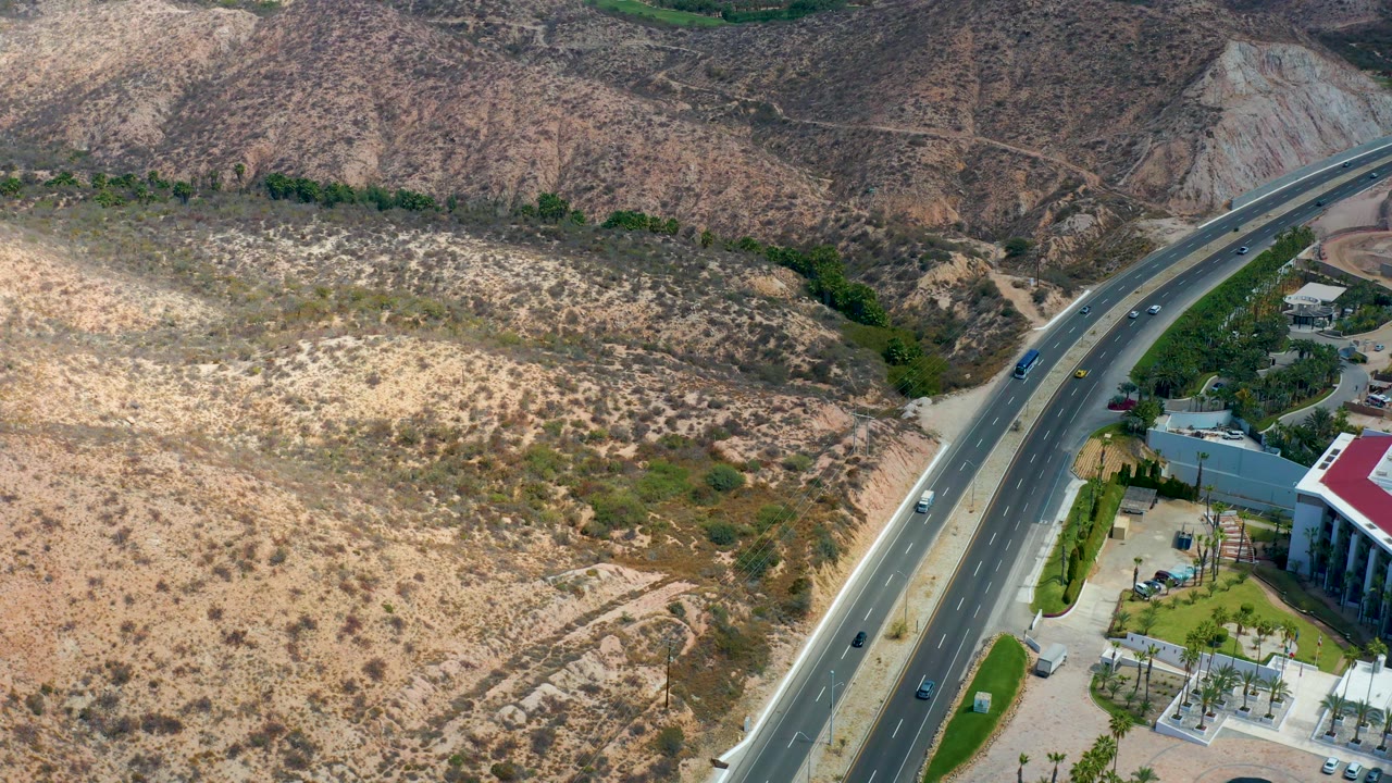 Aerial view at Marquis Los Cabos, Mexico with cars spaced along the divided road, pale rocky hills across the top and left, landscaped greenery below, and no visible people.