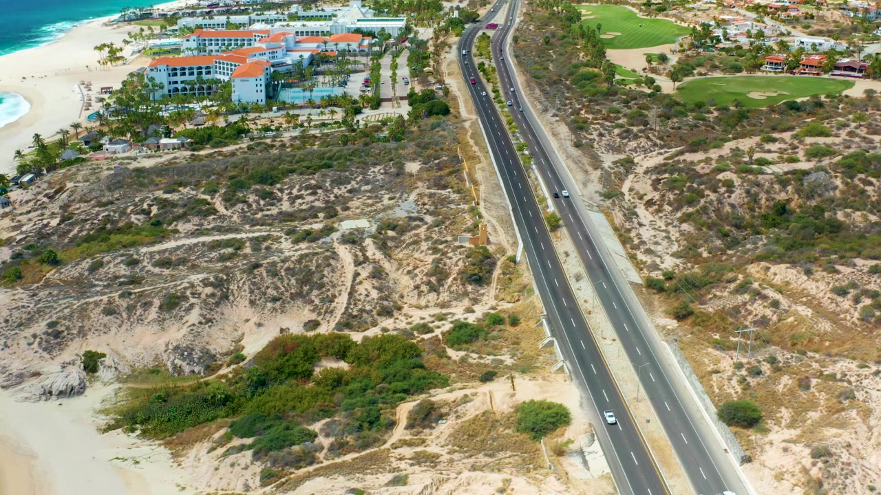 Aerial frame at Marquis Los Cabos showing a divided highway beside sandy dunes, resort buildings near the beach, blue ocean surf at the left edge, and no visible people.