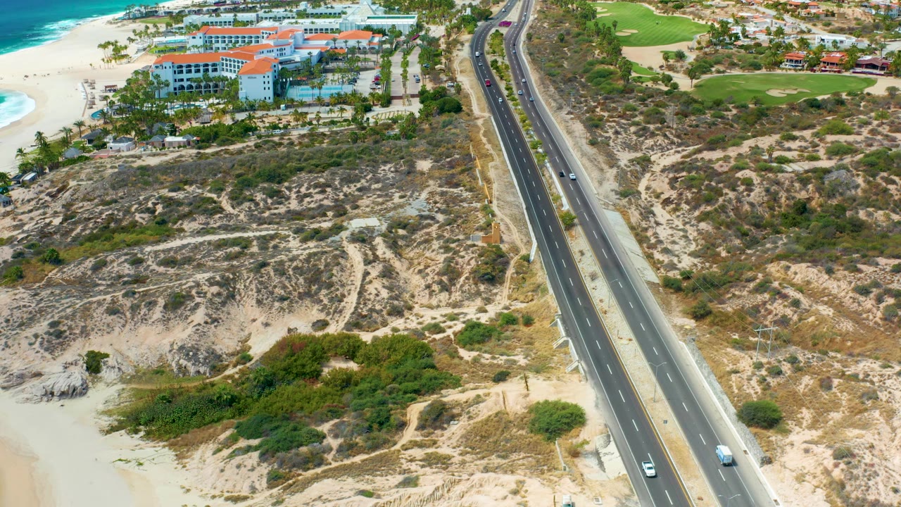 Aerial frame at Marquis Los Cabos showing the highway running diagonally between scrub-covered sand, beachside resort buildings, turquoise surf, green terrain, and no visible people.