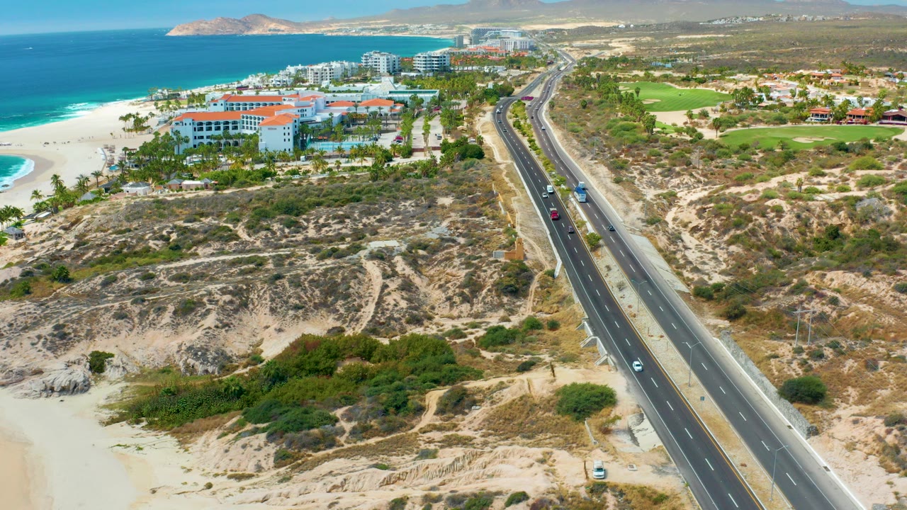 Aerial view at Marquis Los Cabos, Mexico showing a divided highway beside sandy scrub, beach, ocean, resort buildings, green fields, distant hills, and no visible people.