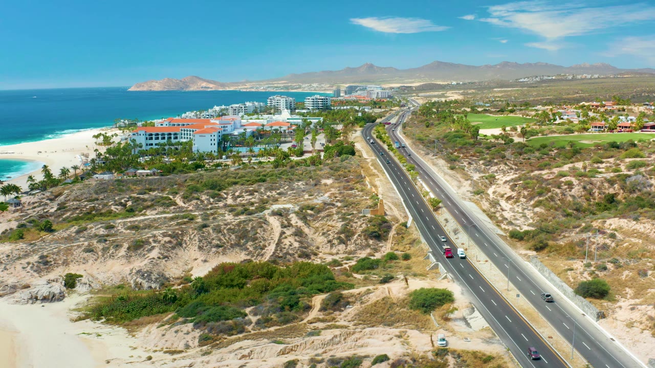 Aerial view at Marquis Los Cabos, Mexico with vehicles spaced along the diagonal highway, sandy beach and blue ocean to the left, white resort buildings ahead, mountains on the horizon, and no visible people.