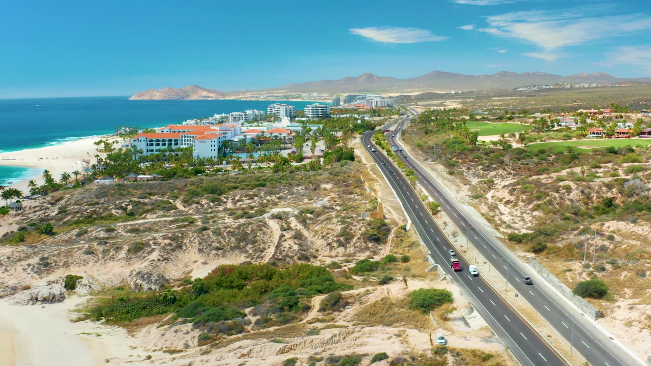 A high aerial frame at Marquis Los Cabos, Mexico shows the highway curving through dry coastal terrain, with a red car and white cars on the road, ocean and beach at left, resort buildings in back, and no visible people.
