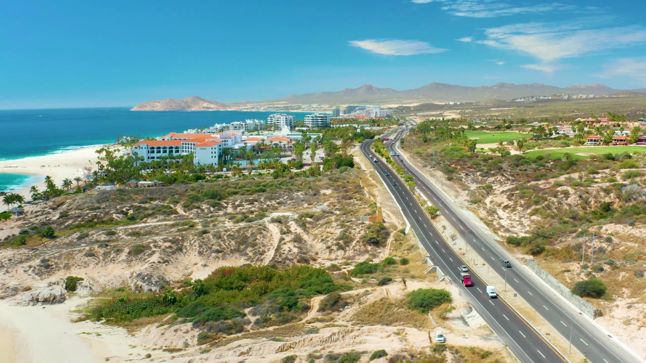At Marquis Los Cabos, Mexico, the frame shows a diagonal highway with several cars, rocky sandy foreground, a strip of beach and blue ocean on the left, resort buildings near the shore, mountains in the distance, and no visible people.