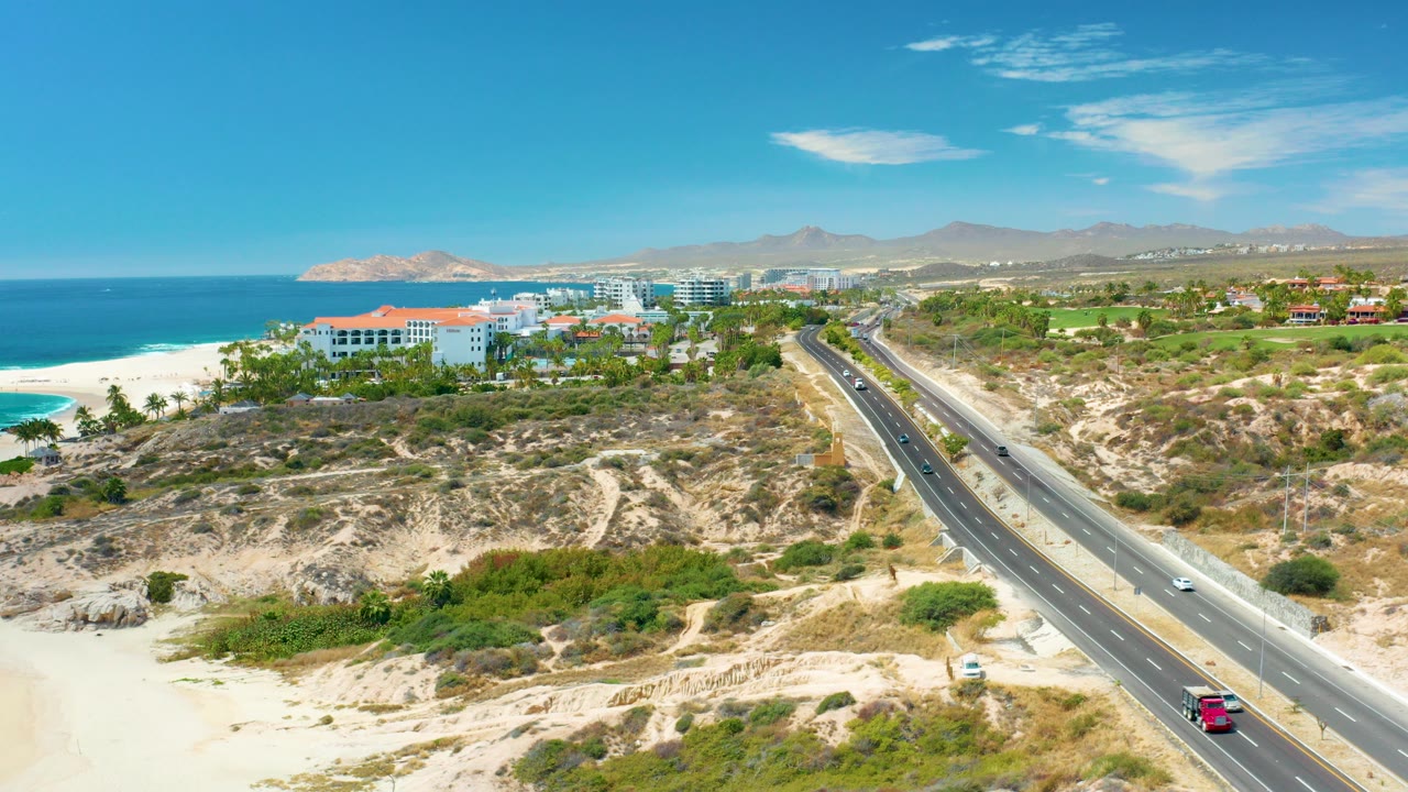 Wide elevated view at Marquis Los Cabos, Mexico with a coastal highway running beside sandy dunes, blue ocean, resort buildings, distant hills, and no visible people.