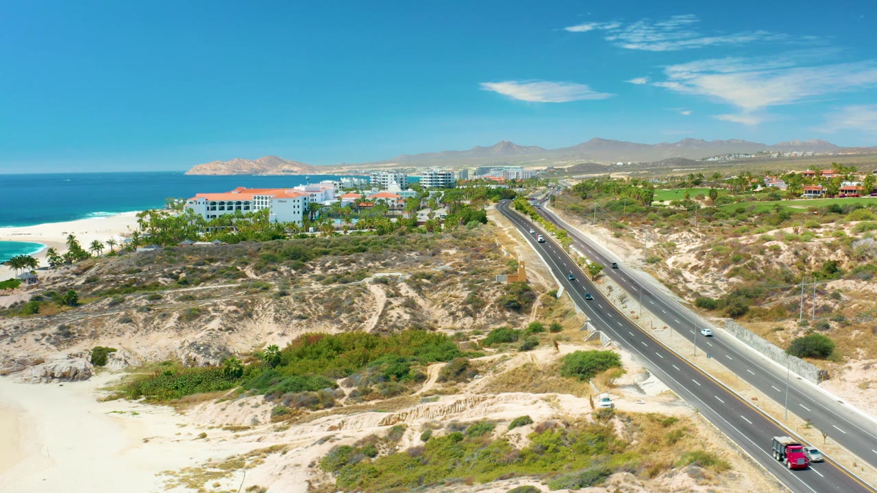At 0:02, the Marquis Los Cabos highway remains centered through sandy dunes, with several vehicles spaced across both directions, ocean to the left, and no visible people.
