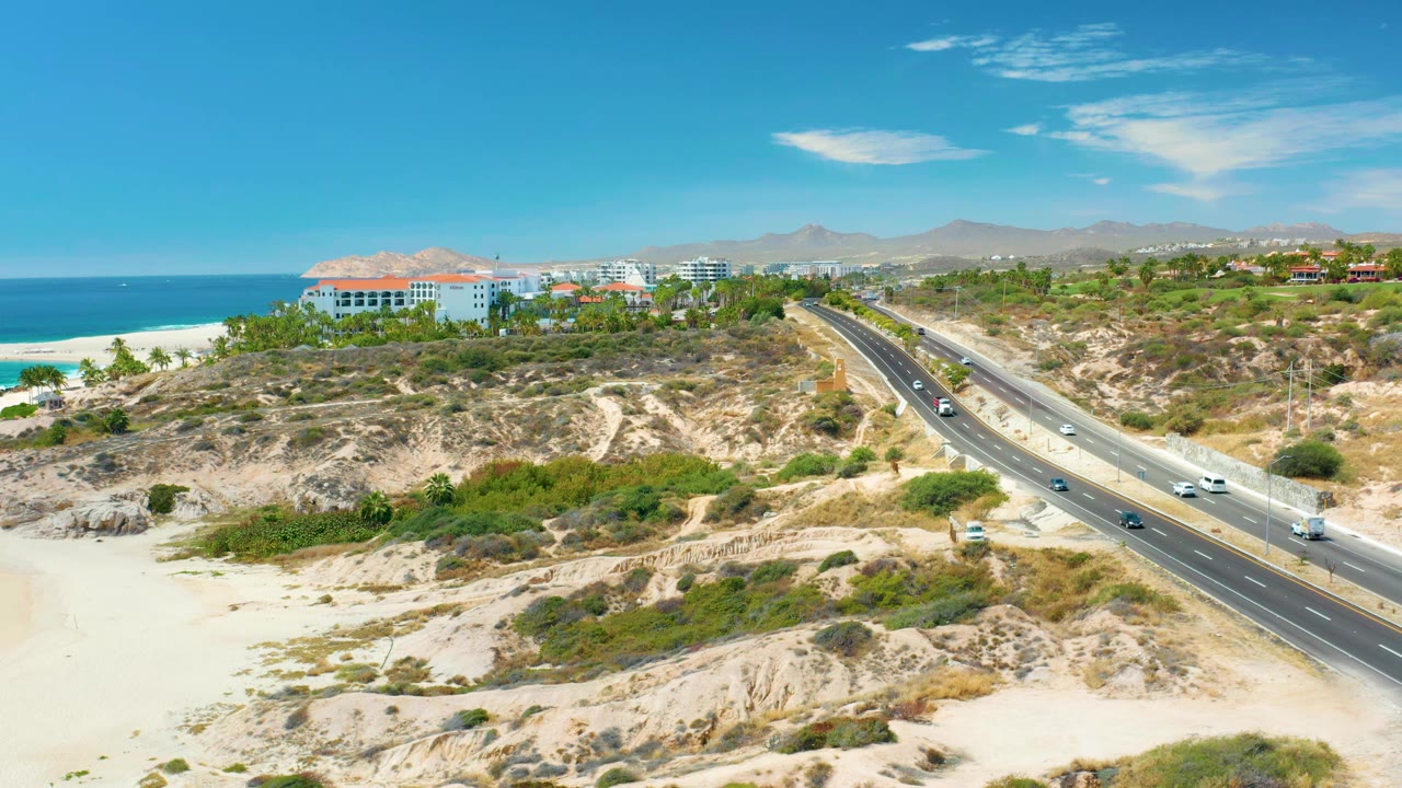 Aerial view one second in shows cars spaced along the divided highway in Marquis Los Cabos, Mexico, with ocean water and pale beach to the left and no visible people.