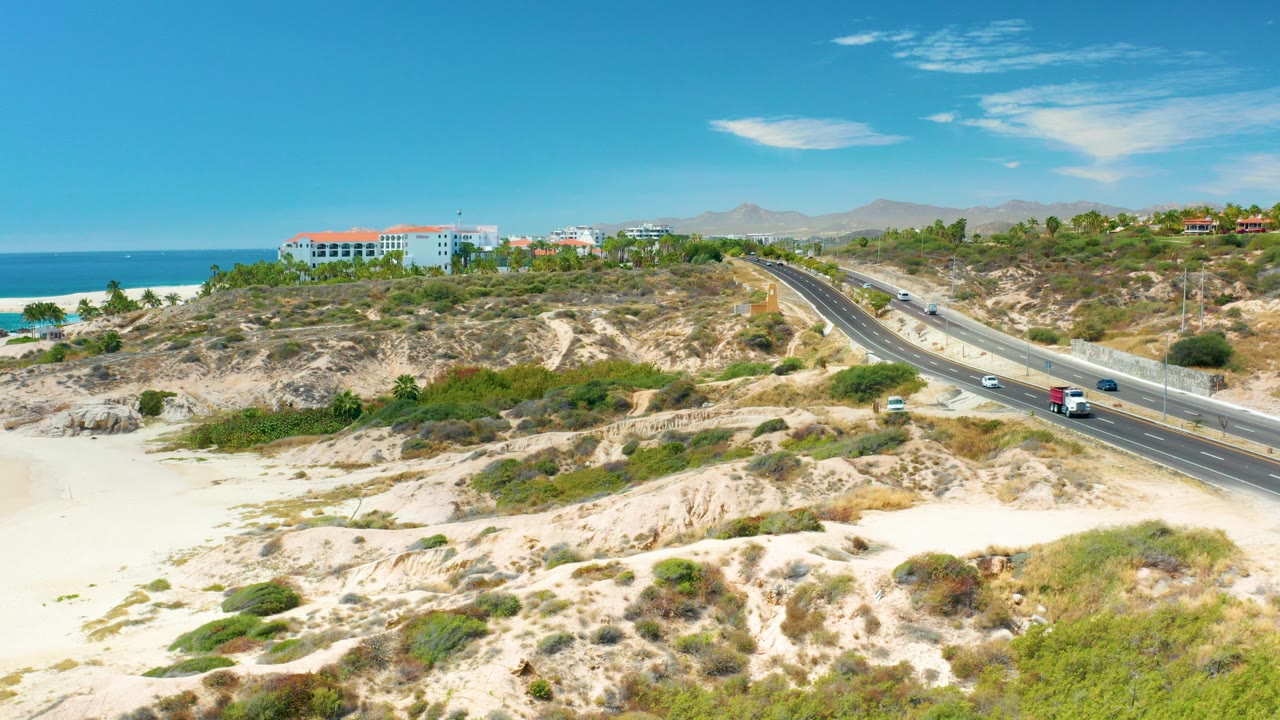 Aerial view at Marquis Los Cabos, Mexico showing sandy dunes, scrub vegetation, a diagonal coastal road with vehicles, beach, ocean, blue sky, and no visible people.