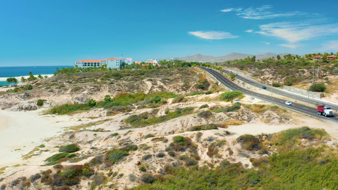 Coastal highway curves past sandy hills with ocean and buildings behind.