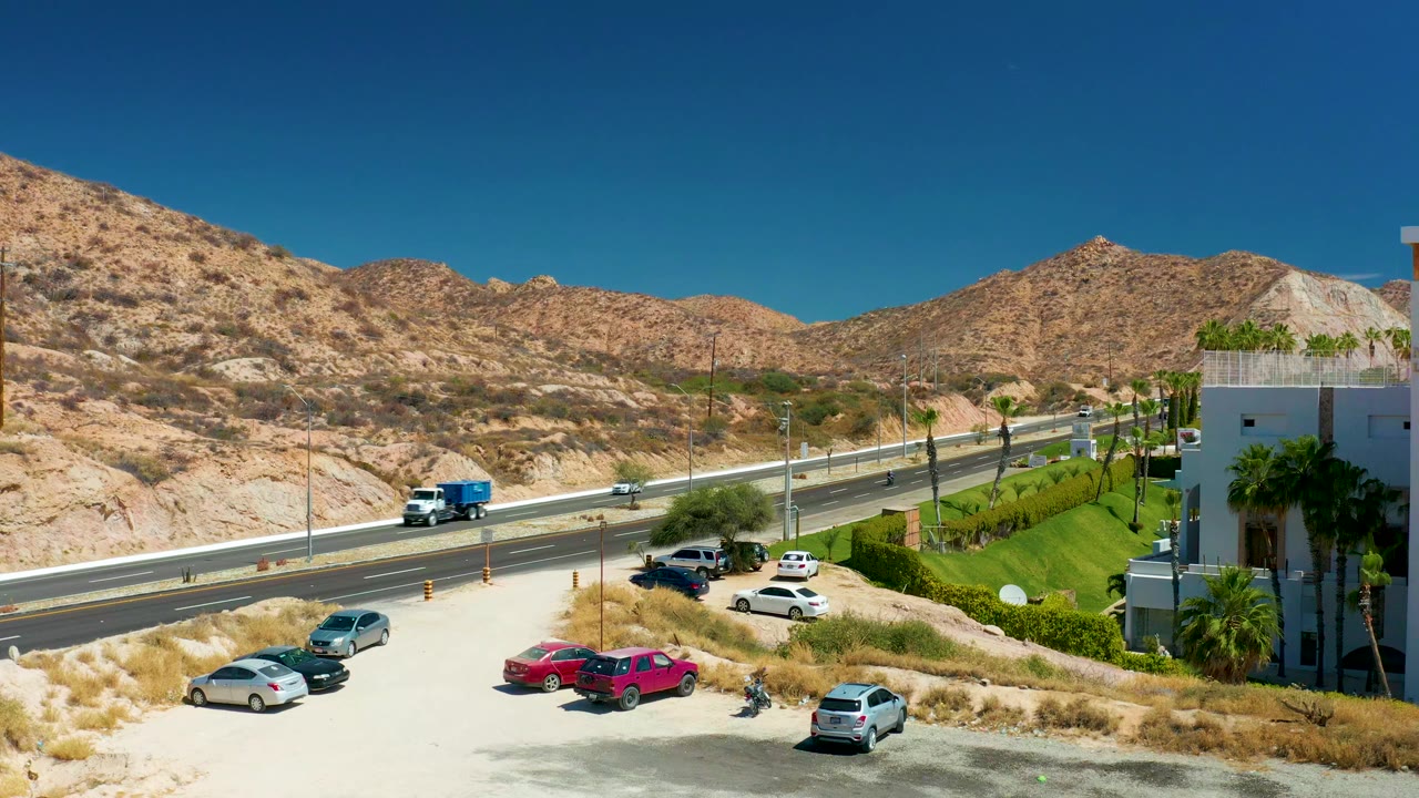 Elevated view at Marquis Los Cabos, Mexico showing a highway, parked cars, tan desert mountains, green landscaping, and a clear blue sky with no visible people.