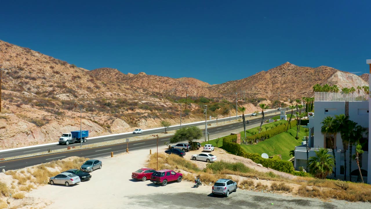 A white cab and blue trailer truck crosses the highway view at Marquis Los Cabos, Mexico with small cars, parked vehicles, rocky desert slopes, and no visible people.