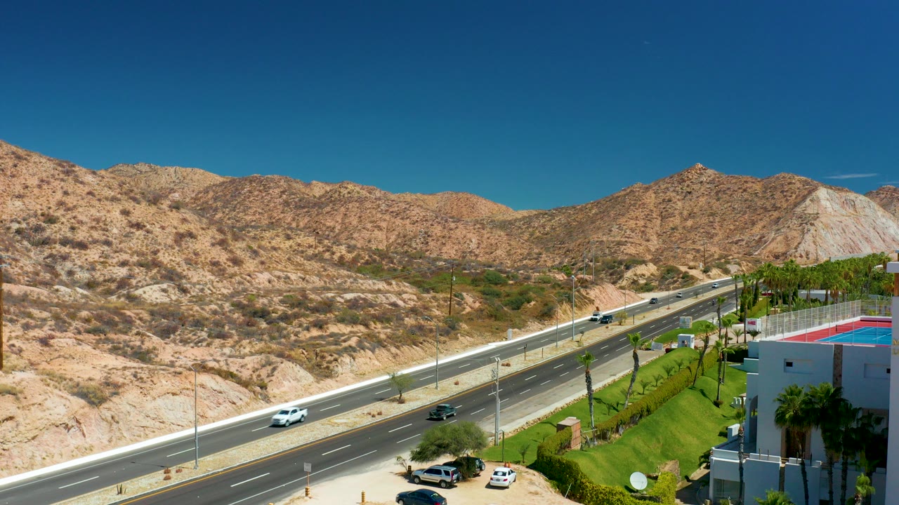 Opening frame of highway traffic in Marquis Los Cabos, Mexico, with tan rocky hills, sparse brush, green landscaping, a red-roofed building edge, and no visible people.