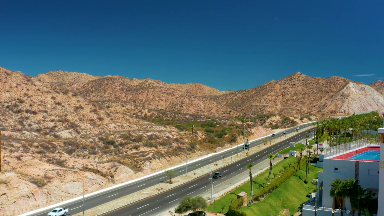 A later highway view in Marquis Los Cabos, Mexico, shows vehicles spaced along the lanes, tan hills across the horizon, green palms and lawn at right, and no visible people.