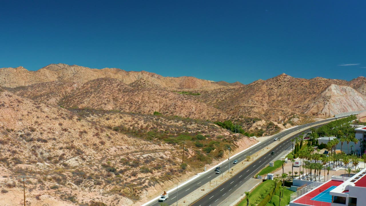 Opening aerial view near Marquis Los Cabos, Mexico, showing a divided highway through dry hills, rocky mountains, blue sky, resort grounds, and no visible people.