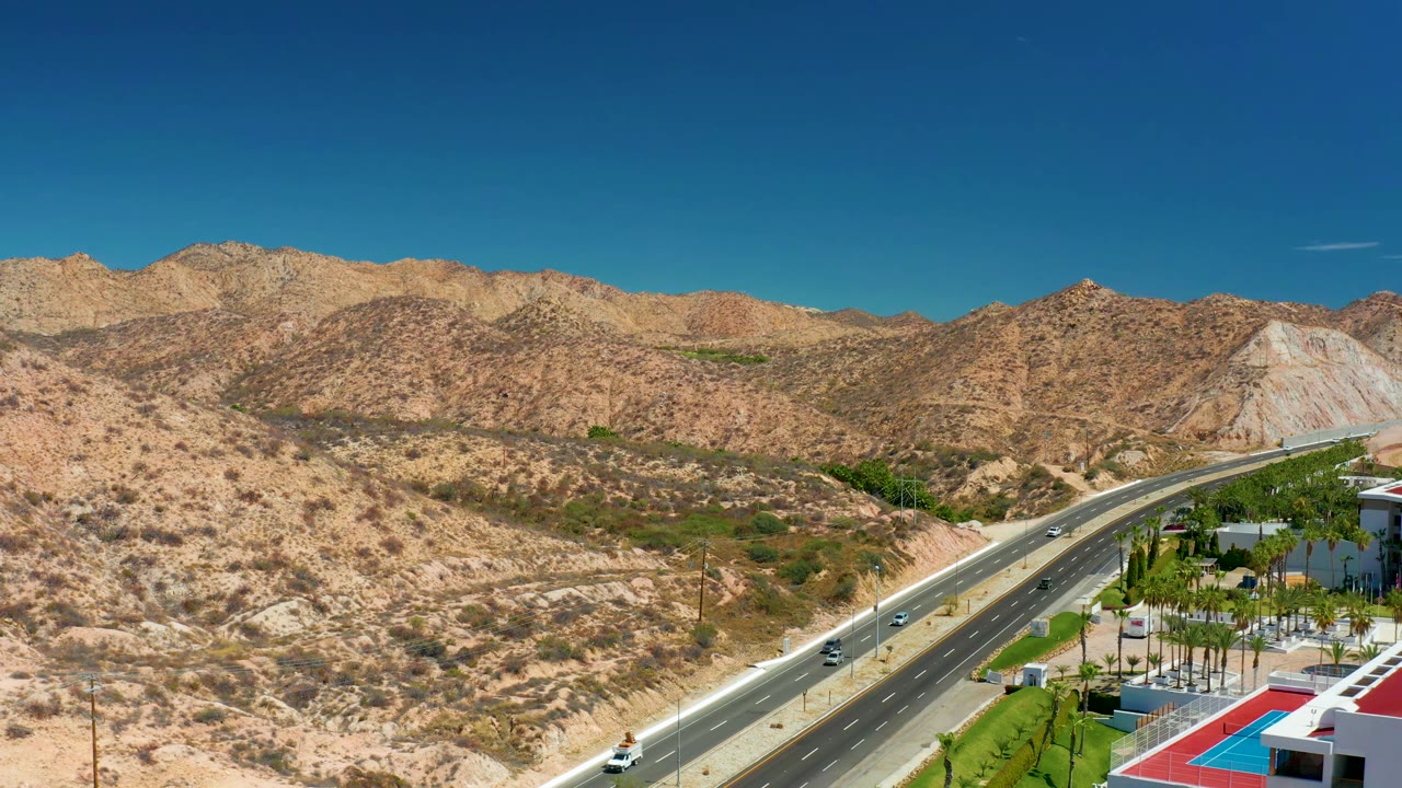 Aerial frame near Marquis Los Cabos, Mexico, with cars spaced along the highway, tan desert slopes, resort pool grounds at lower right, and no visible people.