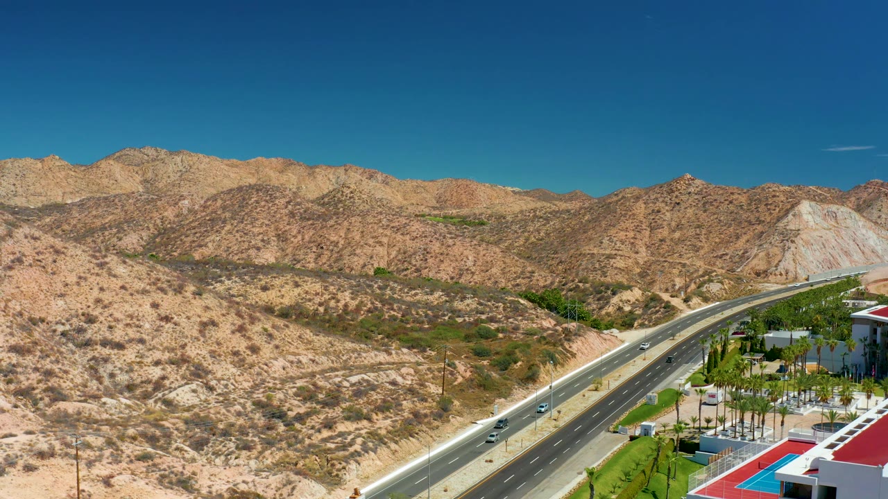 Aerial view near Marquis Los Cabos, Mexico, with the highway angling through arid terrain, several vehicles on the lanes, mountains beyond, and no visible people.