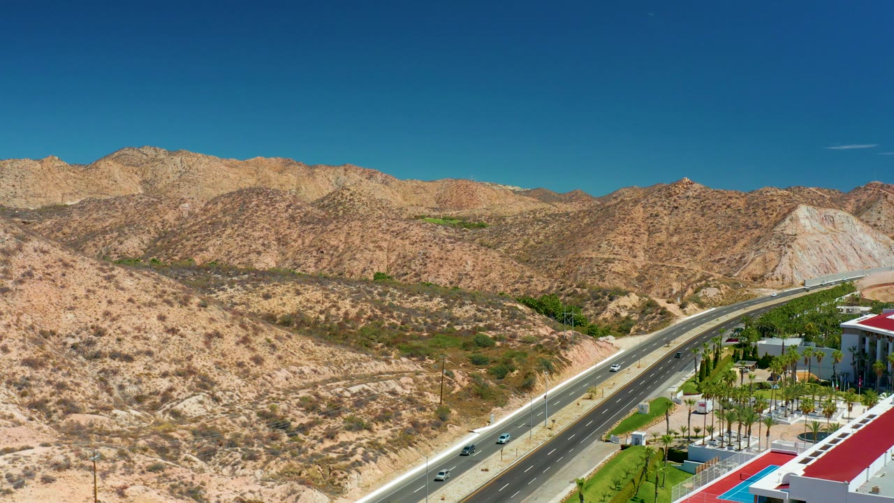 Mid-clip aerial frame near Marquis Los Cabos, Mexico, showing the road curving past scrub-covered hills, resort roofs and pool at lower right, and no visible people.