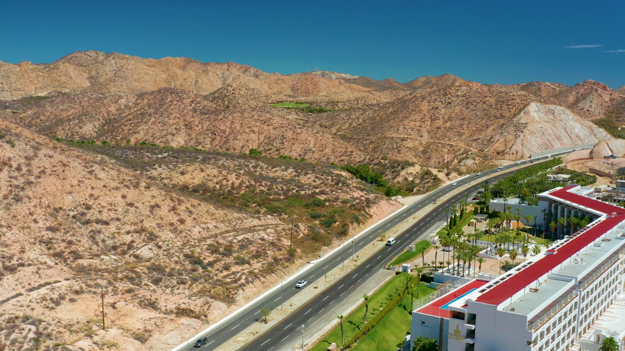 Aerial view near Marquis Los Cabos showing a diagonal highway, dry rocky hills, sparse vegetation, a red-roofed building at lower right, and no visible people.