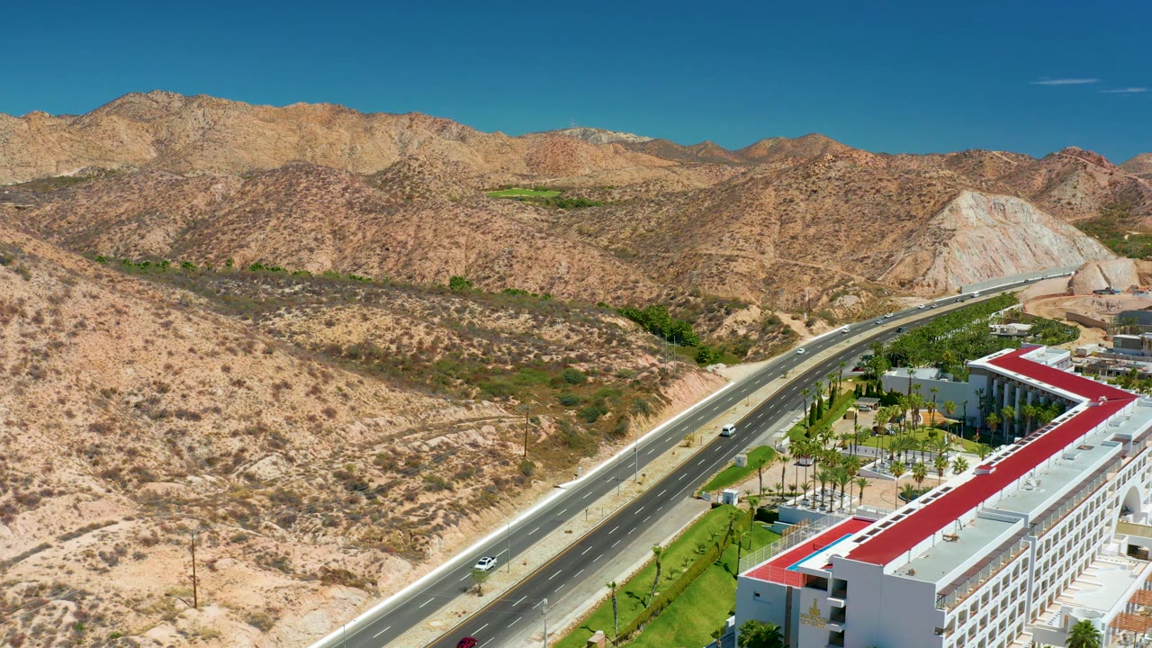 Aerial view near Marquis Los Cabos with highway traffic in the lower half, tan rocky hills across the background, a red-roofed building edge, and no visible people.