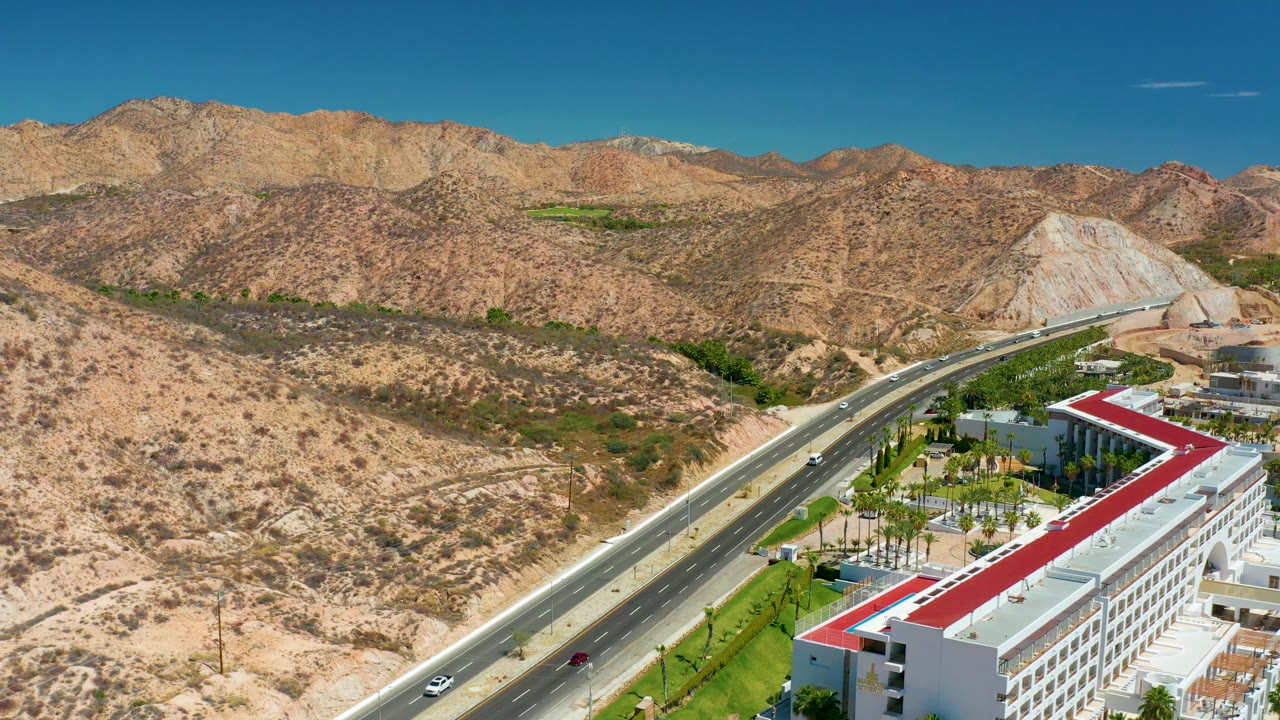 Aerial frame near Marquis Los Cabos showing the highway angled from lower left toward the hills, cars on the road, landscaped greenery near buildings, and no visible people.