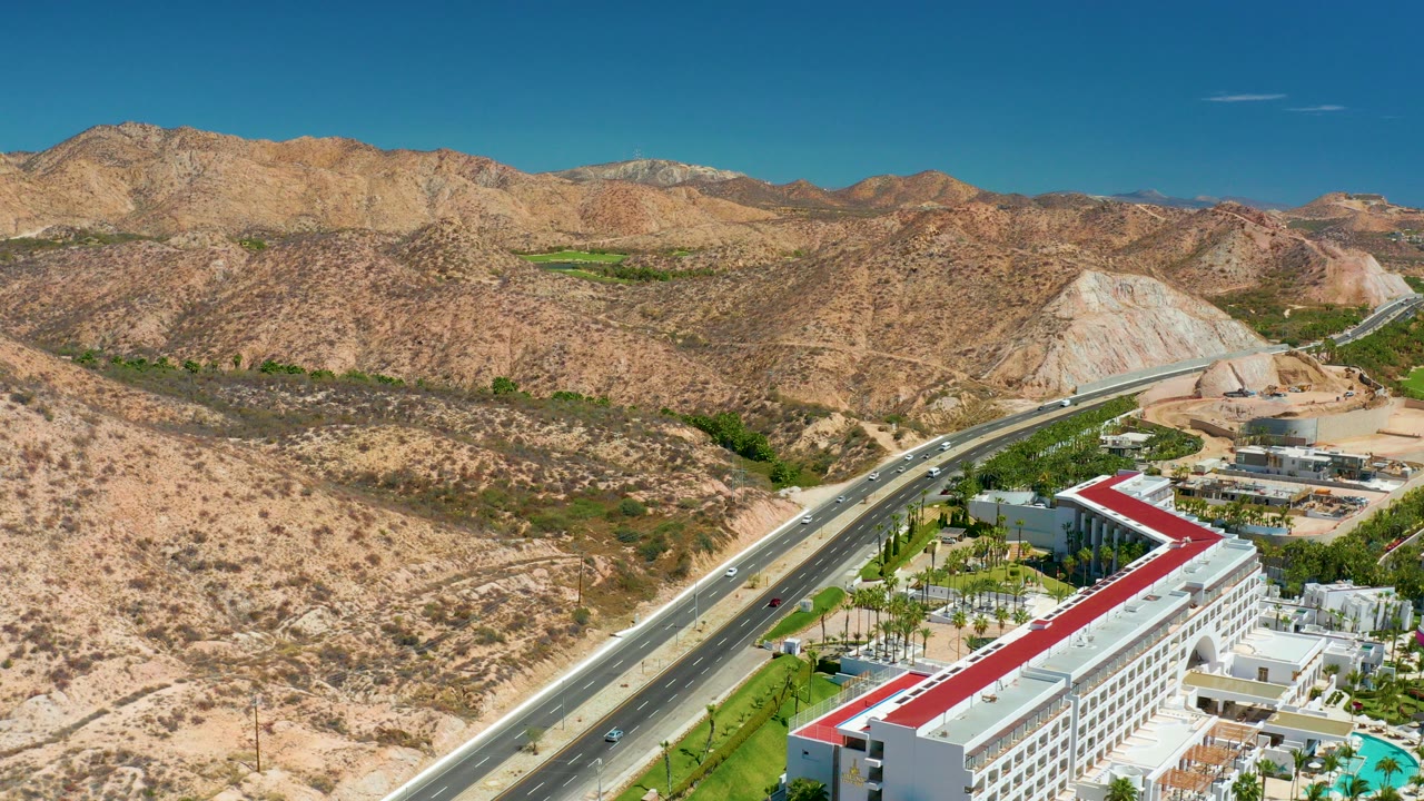 Opening aerial frame over Marquis Los Cabos, Mexico, showing a diagonal highway beside white resort buildings with a red roof, dry hills, distant mountains, and no visible people.