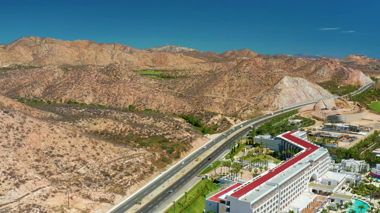 Desert road scene with white buildings, red roof, mountains, and clear sky.