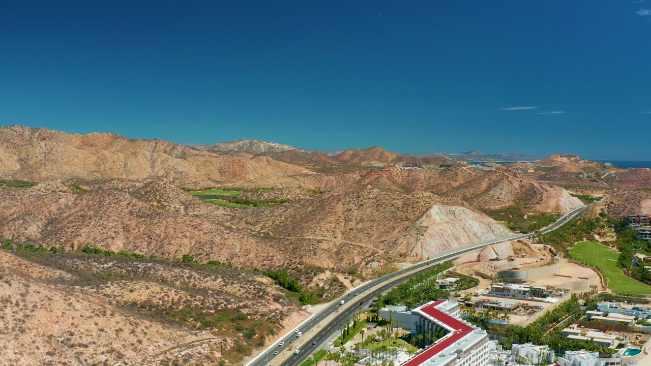 Opening aerial frame at Marquis Los Cabos shows a highway curving through tan hills with low buildings, green landscaping, a red roof edge, distant water, clear sky, and no visible people.
