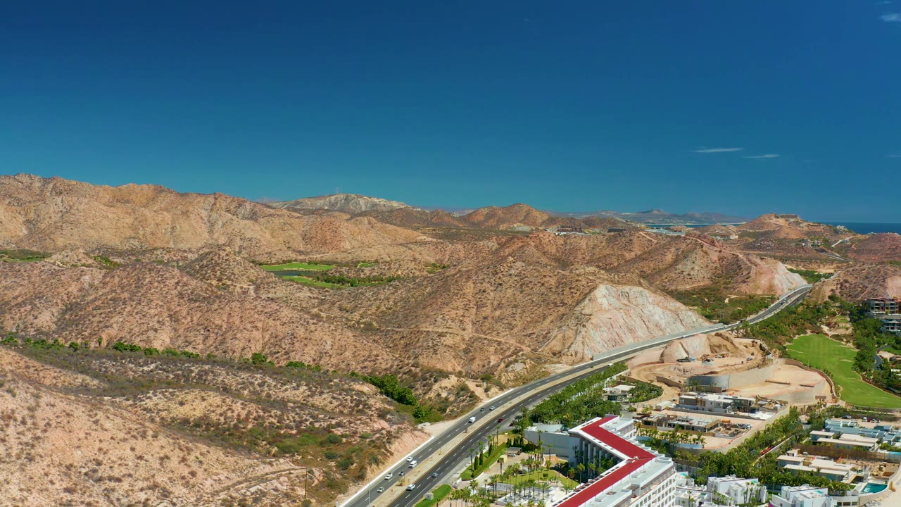 At 0:01, the aerial view holds over Marquis Los Cabos with the divided highway crossing the lower left, tan ridges across the center, green landscaped areas, distant blue water, and no visible people.