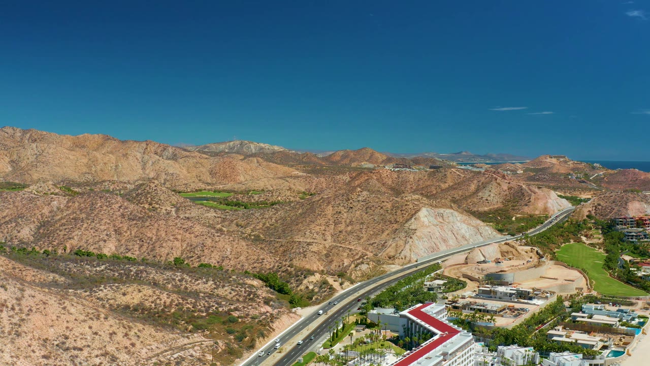 At 0:02, the frame shows small vehicles on a highway below tan hills, with construction-colored ground, low buildings, green course-like landscaping, distant sea, blue sky, and no visible people.