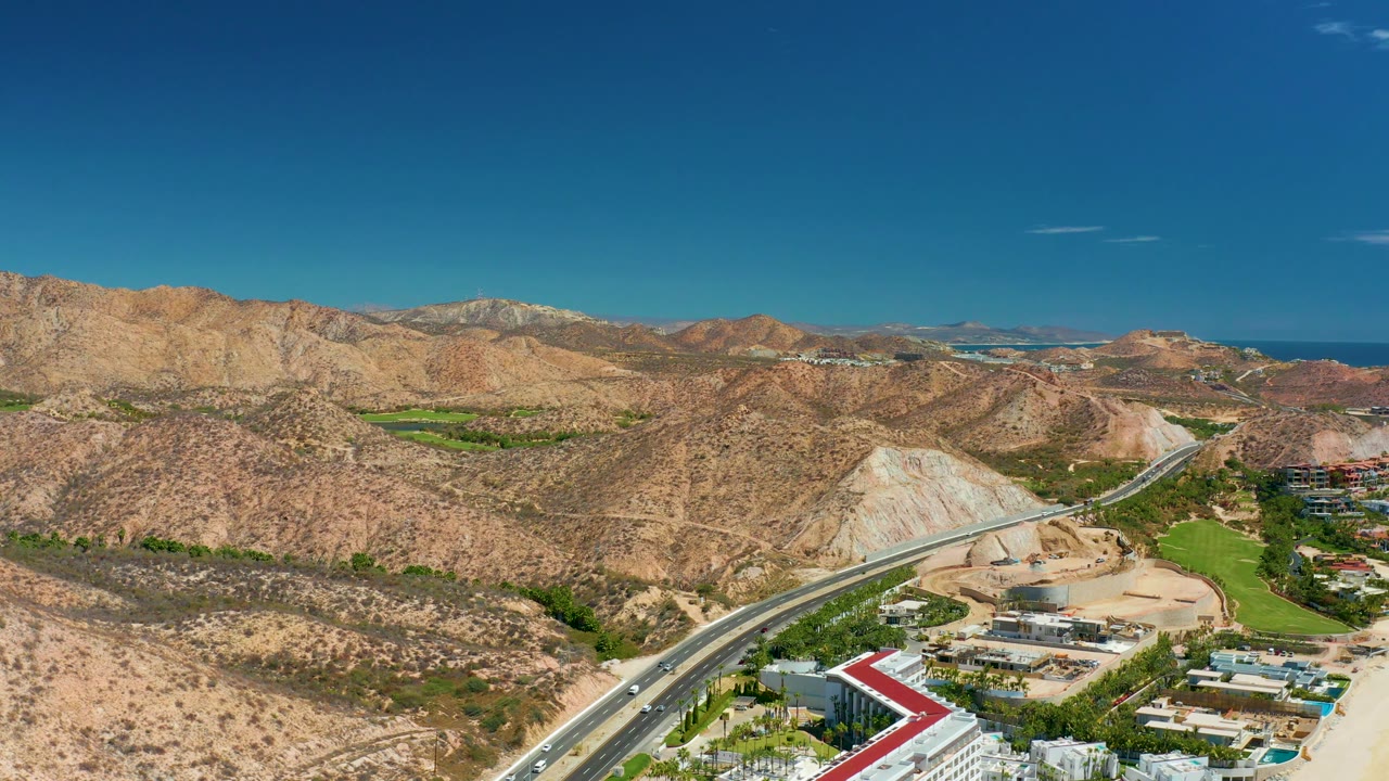 At 0:03, the composition shifts slightly lower over the road corridor, with more foreground buildings and sandy ground visible beneath the dry hills, distant water, and clear sky at Marquis Los Cabos.