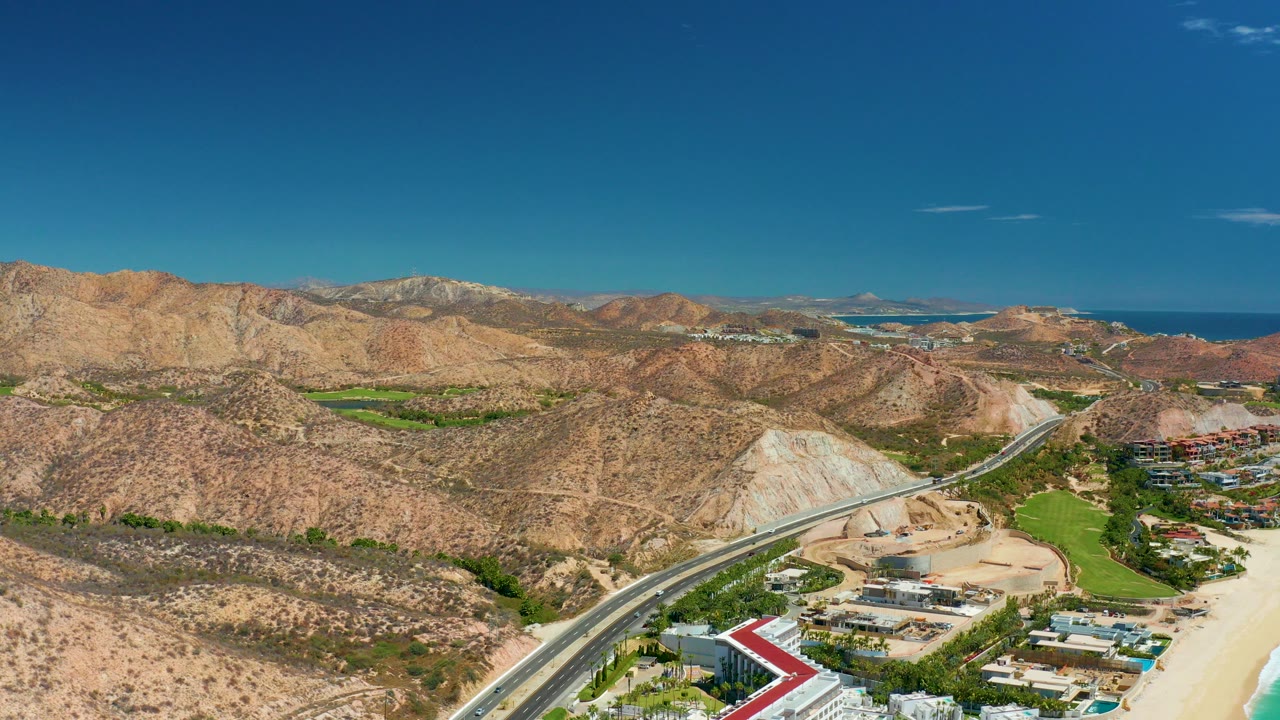 Aerial view near Marquis Los Cabos, Mexico showing a coastal road, sandy beach edge, green fields, arid hills, distant ocean, clear blue sky, and no visible people.