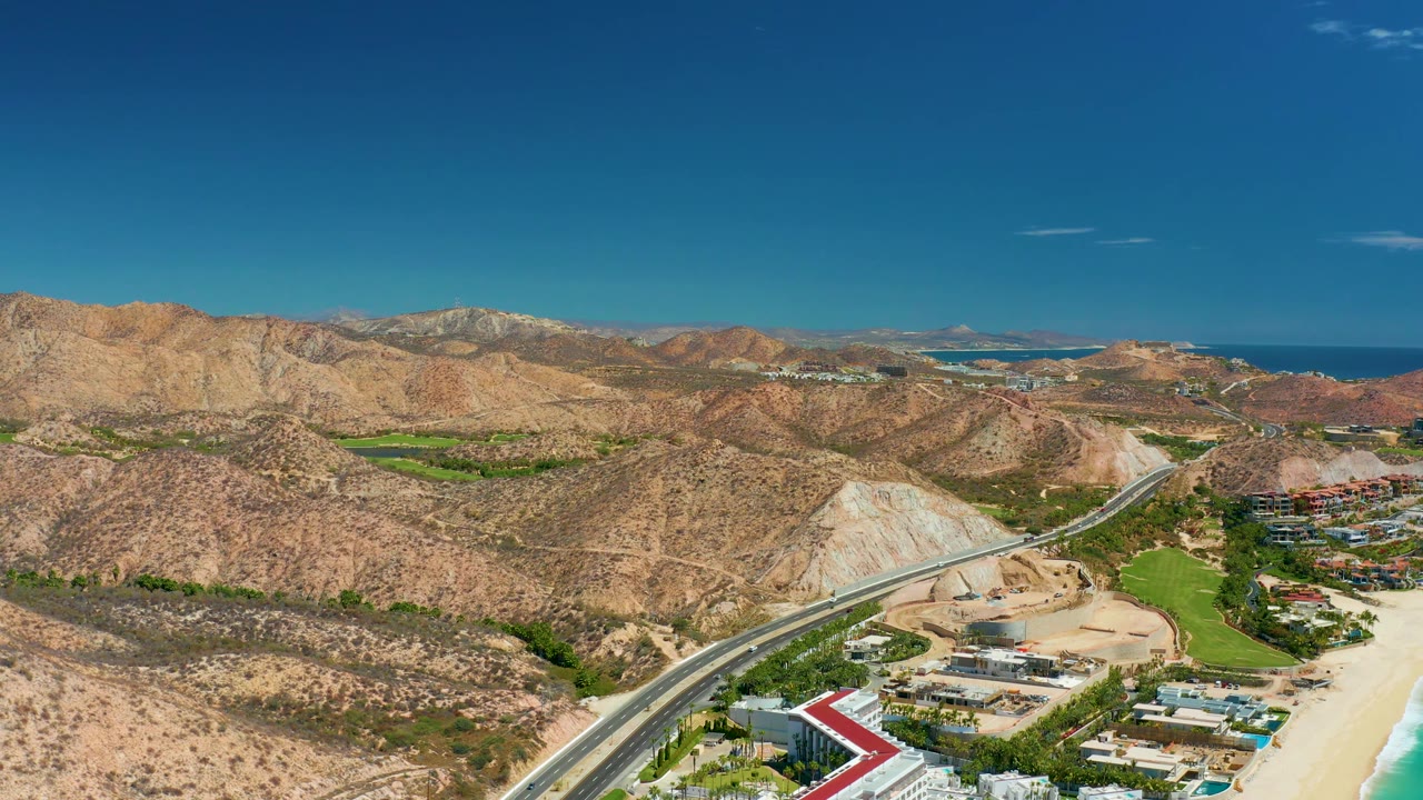 Aerial frame near Marquis Los Cabos, Mexico showing low coastal buildings, a red-roofed structure, sandy beach, turquoise shoreline water, desert hills, distant ocean, and no visible people.