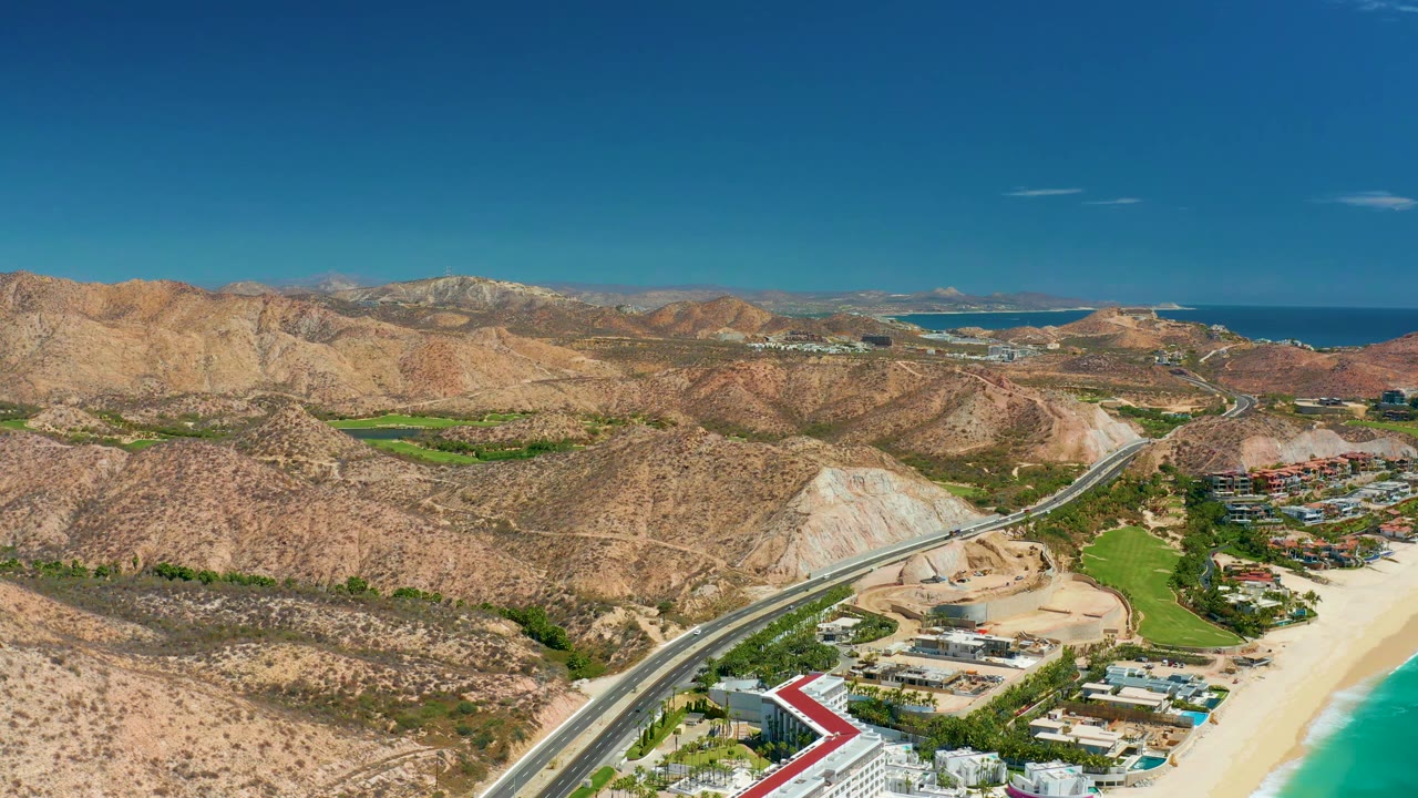 Aerial view in Marquis Los Cabos, Mexico, with the highway crossing the lower frame, resort-like buildings near the beach, dry mountains beyond, ocean on the horizon, and no visible people.