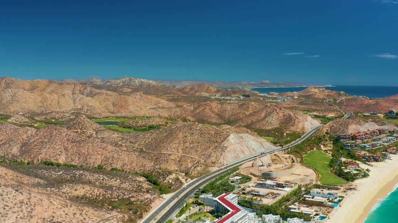 Aerial coastal road curving below desert mountains near a beach.