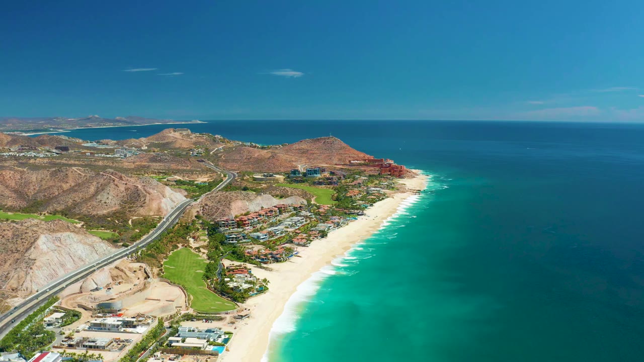 Opening aerial frame over Marquis Los Cabos BCS Mexico shows a sandy beach curving beside turquoise surf, a coastal highway and dry hills to the left, and no visible people.